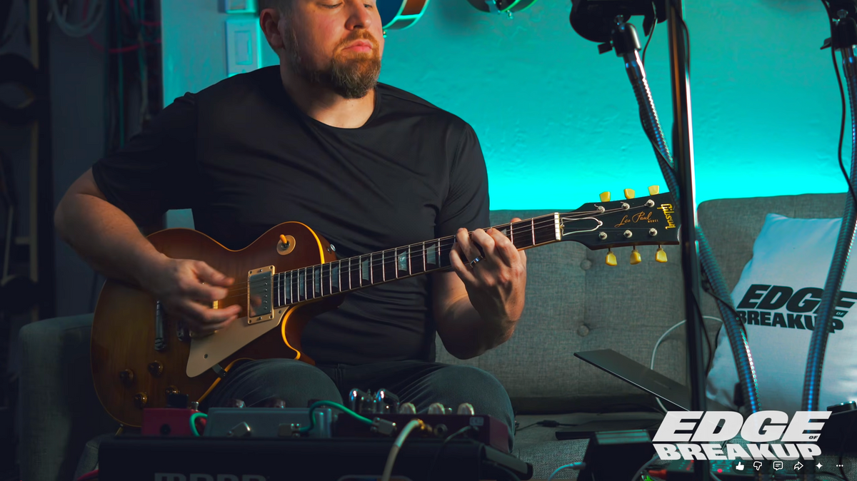 A man with a beard wearing a black t-shirt sits on a gray couch, playing a gold-finish electric guitar. The scene is illuminated by a strong blue-green ambient light, with a microphone stand, effects pedals, and a white pillow with 'EDGE BREAKUP' printed on it in the background.